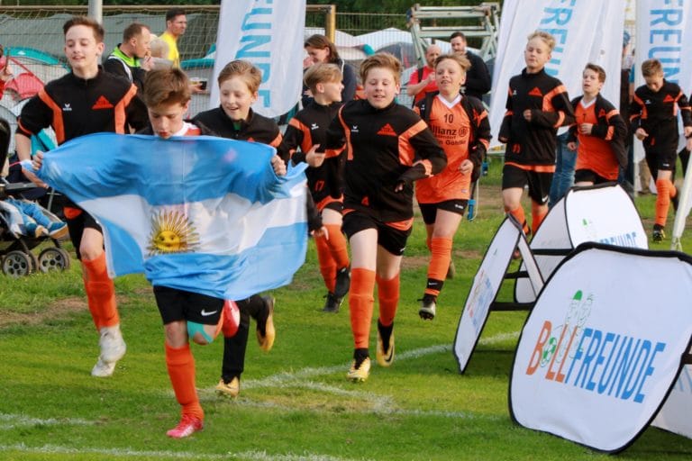 Mehrere jugendliche Fußballer laufen auf einen Fußballplatz ein mit der Landesflagge Argentiniens