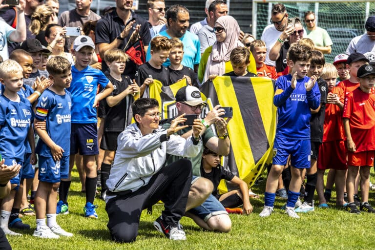 Deux adultes prennent des photos de football avec, en arrière-plan, de nombreux jeunes enfants en tenue de football.